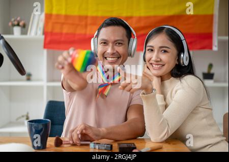 Un uomo gay asiatico attraente e felice con le cuffie che tiene il cuore LGBT nella sua stanza con la sua amica donna. Diversità, omosessuale, gay, amicizia, Foto Stock