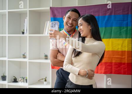 Un'attraente ragazza asiatica sta facendo selfie con il suo amico gay in un salotto con la bandiera LGBT sul muro. Amicizia, diversità, LGBT Foto Stock