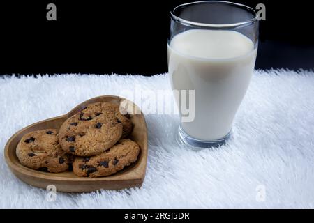Biscotti con gocce di cioccolato su un piatto a forma di cuore con un bicchiere di latte fresco su sfondo nero Foto Stock