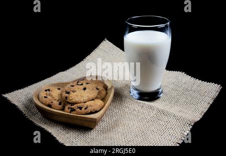 Biscotti con gocce di cioccolato su un piatto a forma di cuore con un bicchiere di latte fresco su sfondo nero Foto Stock
