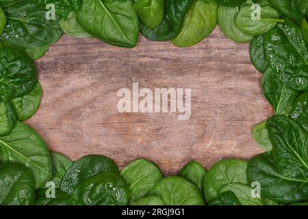 Cornice con bordo, vista piatta dall'alto di una pila di foglie di spinaci leggermente bagnate e fresche, con luce soffusa su uno sfondo rustico in legno con spazio per la copia Foto Stock