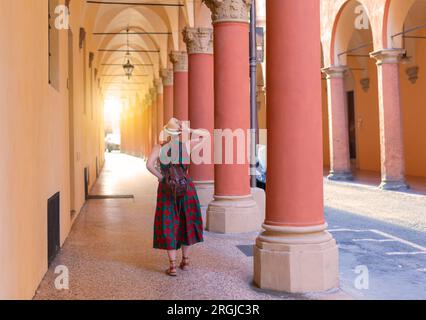 Giovane turista vestita casualmente in piedi con cappello nelle famose gallerie ad arco della città di Bologna in Italia. Bologna è una città studentesca e sede di t Foto Stock