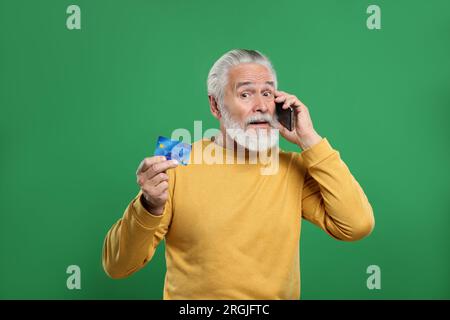 Uomo anziano confuso con carta di credito che parla sullo smartphone su sfondo verde. Attenzione - frode Foto Stock