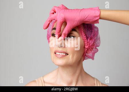 Ritratto di una bella donna sorridente di mezza età che si prepara per le procedure di chirurgia plastica dal medico toccando il tubo in guanti rosa. Foto Stock
