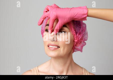 Ritratto di una bella donna sorridente di mezza età che si prepara per le procedure di chirurgia plastica dal medico toccando il tubo in guanti rosa. Foto Stock