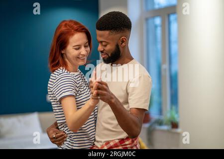 Giovane coppia amante diversa ballare romantica danza in camera da letto moderna con finestre panoramiche. Foto Stock