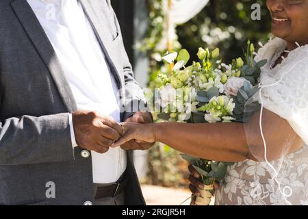 Sezione centrale dello sposo birazziale senior che mette l'anello nuziale sul dito della sposa Foto Stock