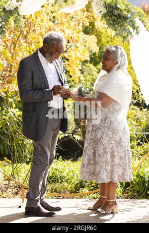 Felice sposo birazziale senior che mette un anello sul dito della sposa alla soleggiata cerimonia nuziale all'aperto Foto Stock