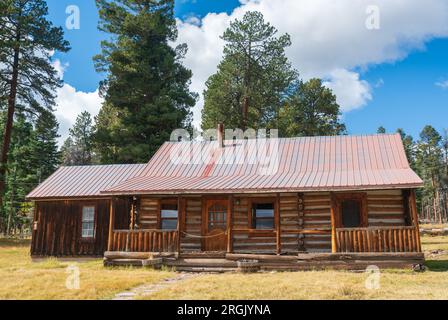 Una capanna di tronchi con un tetto color ruggine e una ringhiera del portico in tronchi è incorniciata da alti pini e un cielo blu, Valles Caldera National Preserve, New Mexico. Foto Stock
