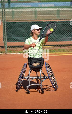 L'uomo pratica tennis su una sedia a rotelle su un campo in terra battuta. Tennis su sedia a rotelle. Foto Stock