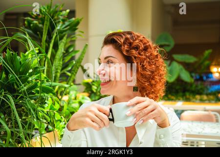 Bella donna alla moda dai capelli rossi beve caffè in un caffè. Foto Stock