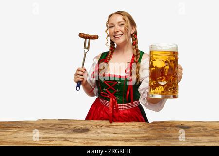 Giovane donna rossa sorridente vestita in Dirndl tradizionale, con un enorme saluto bavarese succoso e appertizzante e una tazza di birra. Concetto di Oktoberfest. Foto Stock