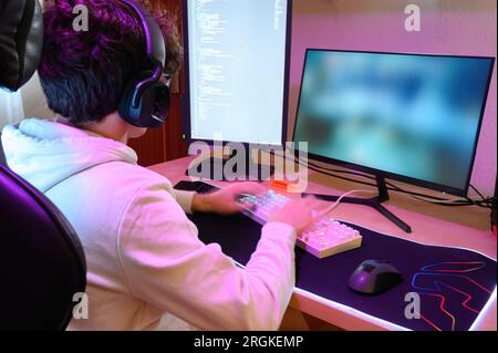 Vista posteriore di un giovane giocatore maschio concentrato in cuffie da vista che guarda lo schermo del monitor mentre si siede al tavolo e si gioca con la tastiera in giro Foto Stock