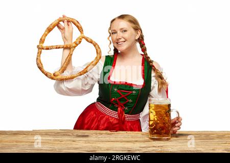 Giovane cameriera, con indosso un tradizionale dirndl bavarese o tedesco, che serve grandi tazze di birra con un enorme pretzel. Concetto Oktoberfest Foto Stock