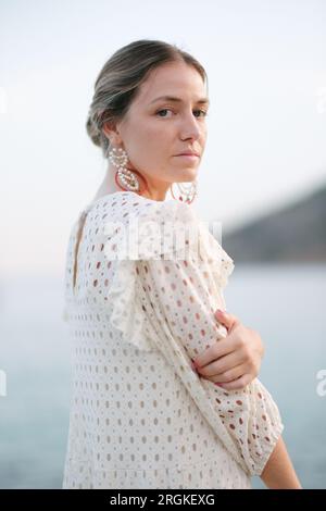Vista laterale di una tranquilla turista adulta in un elegante vestito bianco romantico che tocca il braccio e guarda la macchina fotografica mentre si è in piedi al mare durante l'estate holi Foto Stock