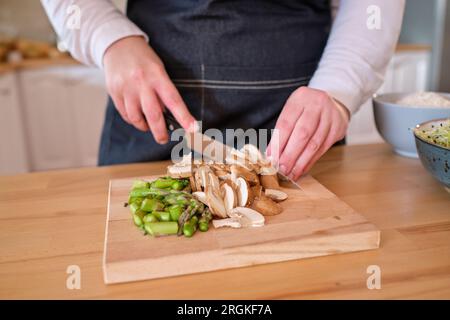 Cuocere nel grembiule e con coltello che taglia funghi crudi e asparagi su tagliere di legno mentre prepari il pranzo vegetariano in cucina Foto Stock