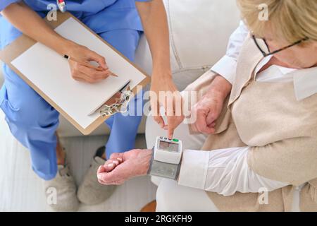 Alto angolo di raccolto medico irriconoscibile con appunti misurazione della pressione sanguigna di una donna anziana con dispositivo sul polso a casa Foto Stock