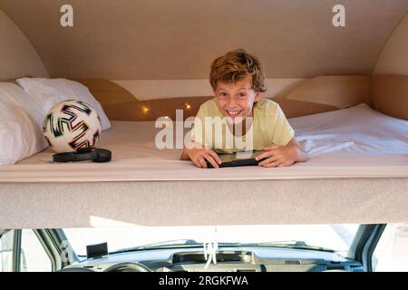 Happy boy con il tablet che sorride e guarda la fotocamera mentre ti rilassi sul letto sul sedile del conducente durante il viaggio in camper Foto Stock