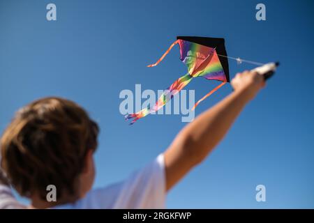 Dal basso verso il basso, vista di un bambino irriconoscibile con aquilone arcobaleno multicolore che vola contro il cielo blu senza nuvole nella soleggiata giornata estiva Foto Stock