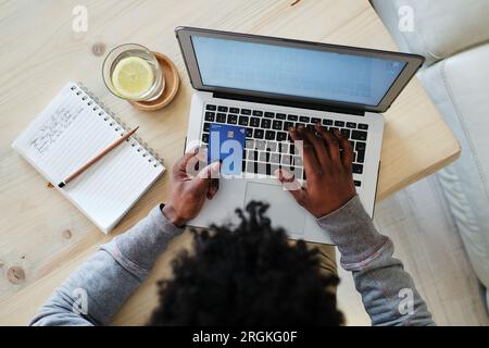 Dall'alto del raccolto irriconoscibile acquirente etnico maschio con carta di plastica che scrive sul netbook mentre facendo l'acquisto a casa Foto Stock
