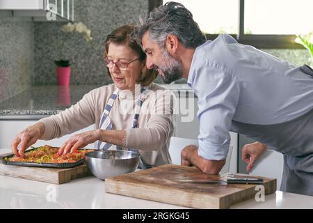 Vista laterale dell'uomo maturo barbuto ispanico con capelli grigi e anziana donna in grembiuli che prepara deliziosi piatti italiani a casa Foto Stock