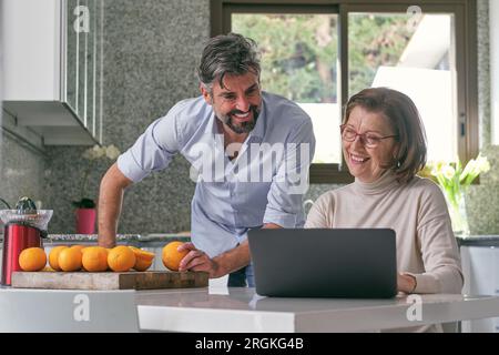 Uomo di mezza età irrasato positivo ispanico, con abiti casual sorridente e in piedi vicino a madre anziana che lavora su un notebook mentre guarda lo schermo insieme Foto Stock