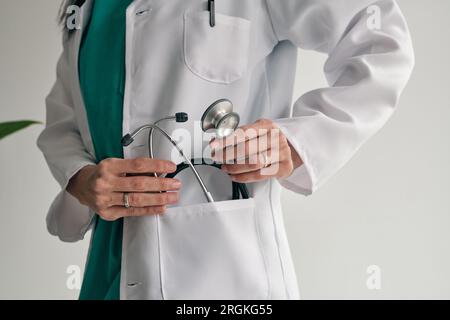 Crop medico femminile irriconoscibile in uniforme medica mettendo stetoscopio in tasca su sfondo chiaro Foto Stock