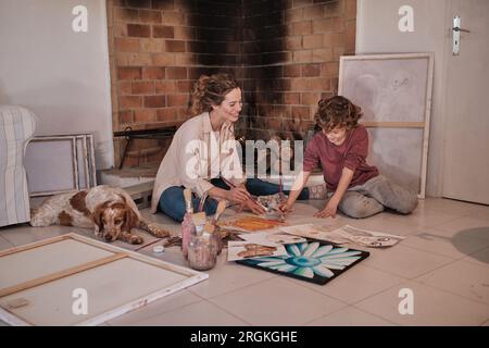 Tutto il corpo di una giovane artista femminile in camicia che si diverte con una bambina dai capelli ricci e dipingono insieme immagini astratte su carta sul pavimento di una casa di campagna Foto Stock