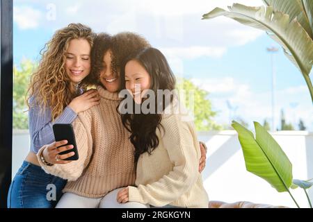 Contenuti giovani donne diverse con maglioni seduti l'uno vicino all'altro e abbracciati mentre si scatta un autoritratto sul telefono cellulare Foto Stock