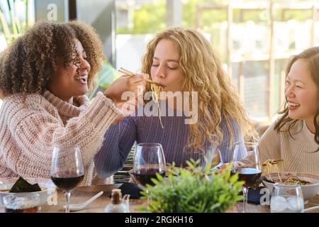 Un gruppo di amici che ridono si divertono mangiando ramen con le bacchette e bevendo vino al ristorante Foto Stock