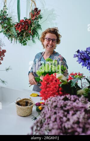 Fiorista adulta positiva in occhiali e grembiule in piedi vicino al bancone con bouquet di trachelio verde circondato da fiori fioriti in sho floreale Foto Stock