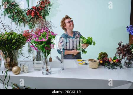 Fiorista adulta positiva con trachelio verde che organizza il bouquet mentre si trova vicino al bancone con fiori assortiti in vasi di vetro Foto Stock