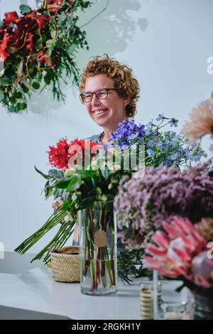 Fiorista adulta con un mazzo di fiori freschi in piedi vicino al bancone con piante in fiore assortite in vasi trasparenti nel negozio di fiori Foto Stock