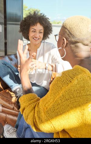 Amiche afro-americane positive che sorridono e si guardano mentre si siedono vicino alla finestra al caffè Foto Stock