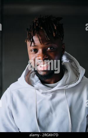 Uomo afro-americano sicuro ed elegante con cappuccio che guarda la fotocamera con espressione positiva del volto mentre si trova in piedi su uno sfondo scuro nella stanza Foto Stock