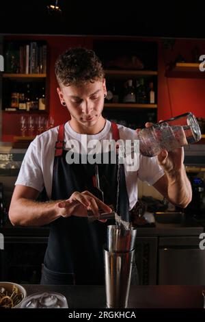 Giovane barista maschio con orecchini in piedi vicino al bancone e versare vodka dalla bottiglia e sciroppo da jigger nell'agitatore ciabattino in metallo durante la preparazione Foto Stock