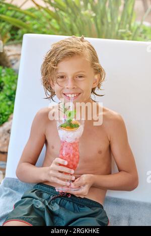 Un giovane preadolescente seduto sulla sedia e guardando la macchina fotografica mentre sorseggia un cocktail fresco con un gelato al sole all'aperto Foto Stock