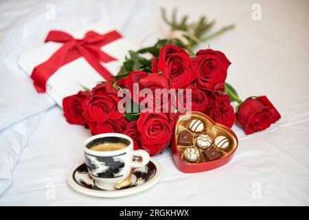 Dall'alto di composizione di mazzo di rose rosse e tazza di caffè fresco aromatico con scatola di cioccolatini a forma di cuore collocata su un morbido letto bianco dell'hotel Foto Stock