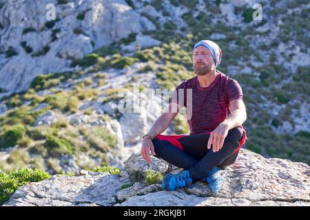 Escursionista maschio barbato seduto su una montagna rocciosa e rilassato dopo il trekking negli altopiani durante le vacanze estive Foto Stock