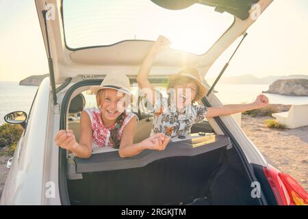 Due bambini in cappelli di paglia seduti sui sedili posteriori di un'automobile che attacca le loro teste fuori del bagagliaio facendo gesti di vittoria con le braccia sollevate e. Foto Stock