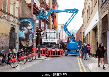 Strada chiusa bloccata da un camion da costruzione con gru e veicoli raccoglitori di ciliegie murale donna nera Spitalfields East London E1 England KATHY DEWITT Foto Stock