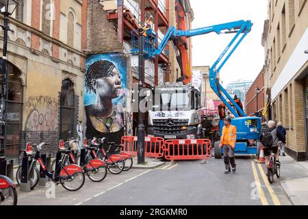 Strada chiusa bloccata da camion da costruzione con gru e veicoli cherrypicker donna nera murale Spitalfields East London E1 UK England KATHY DEWITT Foto Stock