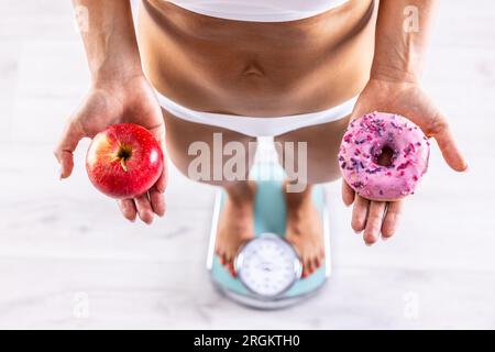 Donna sportiva con un corpo perfetto che misura il peso in scala e regge una ciambella e una mela rossa. Concetto di stile di vita sano o malsano. Foto Stock