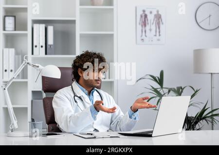 Giovane medico indiano scontento e serio all'interno dell'ufficio che parla da remoto consultando i pazienti utilizzando un computer portatile per riunioni a distanza. Foto Stock