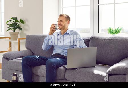 Uomo felice che si prende una pausa durante il lavoro, seduto sul divano con il suo portatile e bevendo un tè caldo Foto Stock