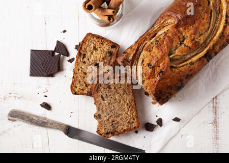Pane alla banana umido a fette con avena e pezzi di cioccolato fondente su un tavolo di legno bianco Foto Stock