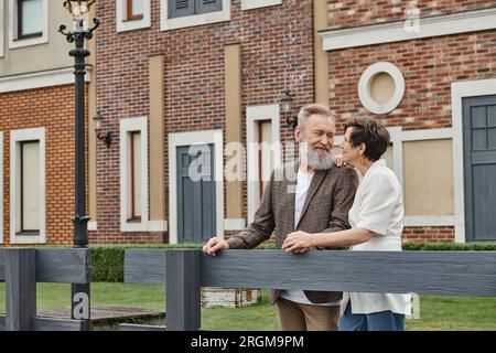 felice coppia anziana, uomo e donna in piedi vicino alla recinzione accanto alla casa, guardarsi l'un l'altro, romanticismo Foto Stock