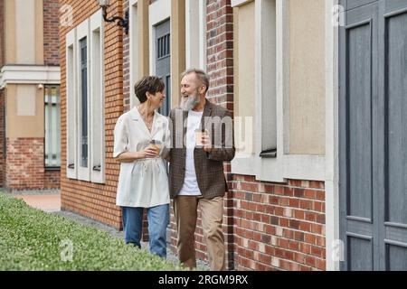 felice coppia anziana che tiene un caffè da andare, camminando vicino all'edificio, uomo e donna anziani, risate Foto Stock