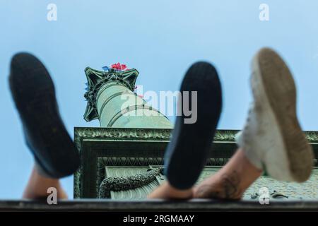 Parigi, Francia, 2018. La colonna di luglio decorata con bandiere francesi per il giorno della Bastiglia, vista tra le scarpe di due uomini seduti sul monumento Foto Stock