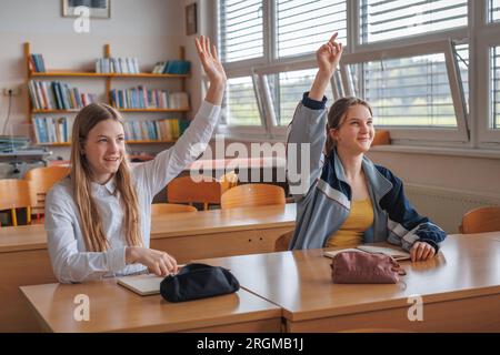 Due adolescenti studentesse alzano le mani per rispondere alla domanda in classe. Concetto di ritorno a scuola. Foto Stock